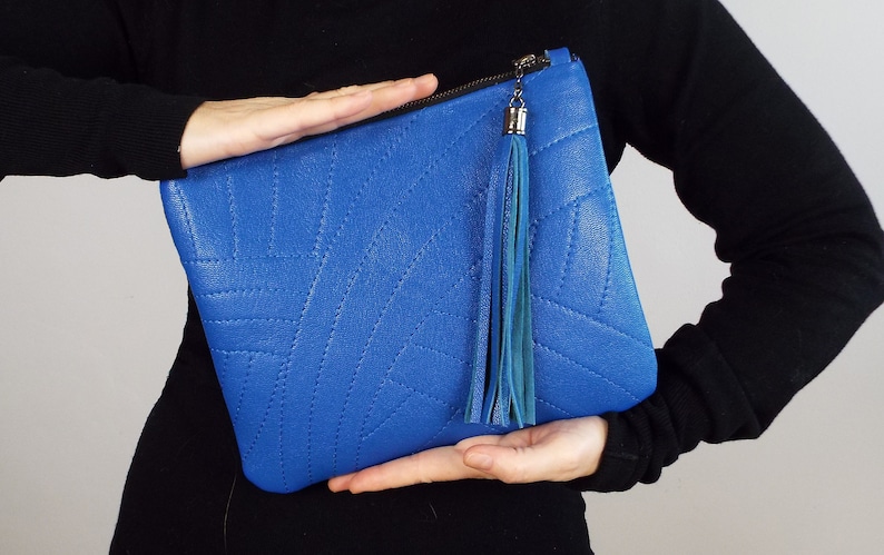 May include: A vibrant blue clutch bag with a stitched geometric design. It features a zip closure and a long, teal tassel. The bag is set against a black background.