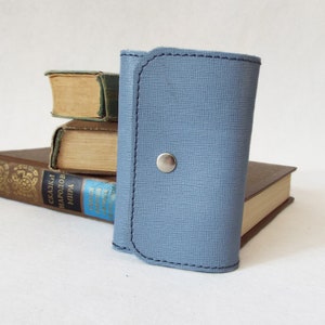 May include: A light blue leather key holder with a silver snap closure. The key holder is sitting on top of a stack of books.