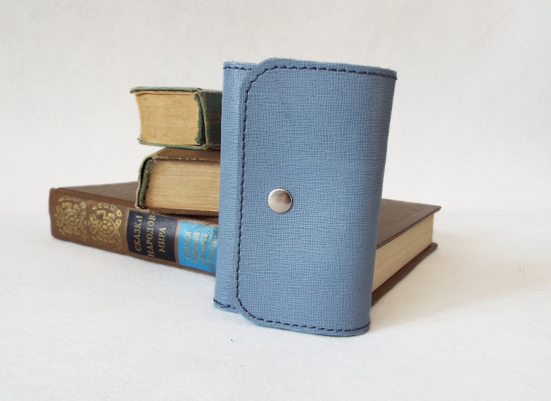 May include: A light blue leather key holder with a silver snap closure. The key holder is sitting on top of a stack of books.
