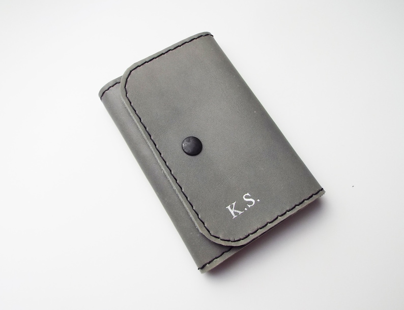 May include: A gray leather wallet with a black button and white stitching. The wallet has the initials "K.S." embossed on the front.