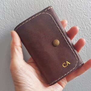 K&ouml;nnte beinhalten: Eine braune Lederb&ouml;rse mit einem goldenen Knopf und den Initialen "CA" in Gold gepr&auml;gt.