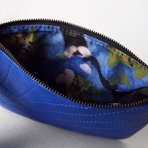 May include: A royal blue quilted leather clutch bag with a black zip and a wrist strap. The interior lining has a colourful floral pattern with blue, yellow, and pink hues. The bag is open, showing the patterned lining.