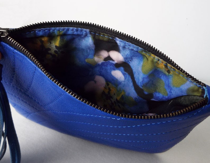 May include: A royal blue quilted leather clutch bag with a black zip and a wrist strap. The interior lining has a colourful floral pattern with blue, yellow, and pink hues. The bag is open, showing the patterned lining.