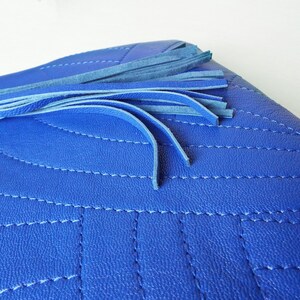 May include: A close-up of a vibrant blue clutch bag featuring a quilted design and a tassel. The bag has a silver-toned metal detail and is made of a textured material. The tassel is made of multiple blue strands.