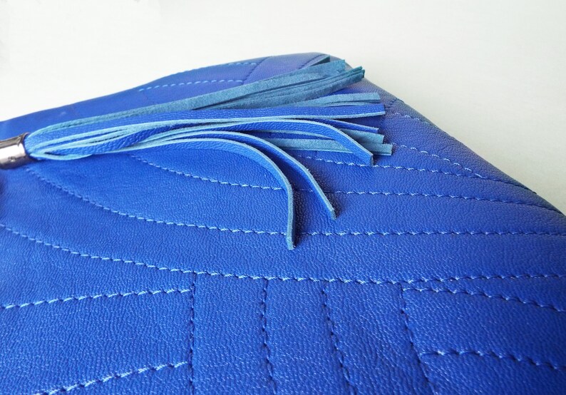 May include: A close-up of a vibrant blue clutch bag featuring a quilted design and a tassel. The bag has a silver-toned metal detail and is made of a textured material. The tassel is made of multiple blue strands.