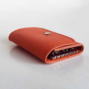 K&ouml;nnte beinhalten: Ein leuchtend orangefarbener Schl&uuml;sselanh&auml;nger aus Leder mit einem Schl&uuml;sselring aus Metall und einem Druckknopfverschluss.
