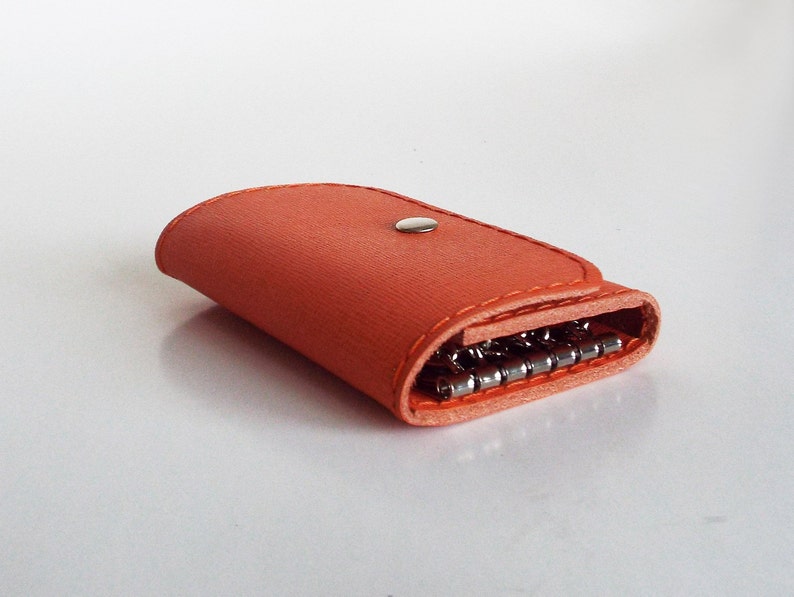 K&ouml;nnte beinhalten: Ein leuchtend orangefarbener Schl&uuml;sselanh&auml;nger aus Leder mit einem Schl&uuml;sselring aus Metall und einem Druckknopfverschluss.