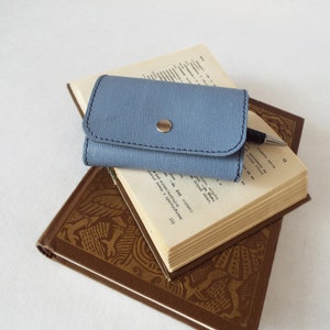 May include: A blue leather key holder with a silver button closure, resting on top of two books. The top book has a pen on it.
