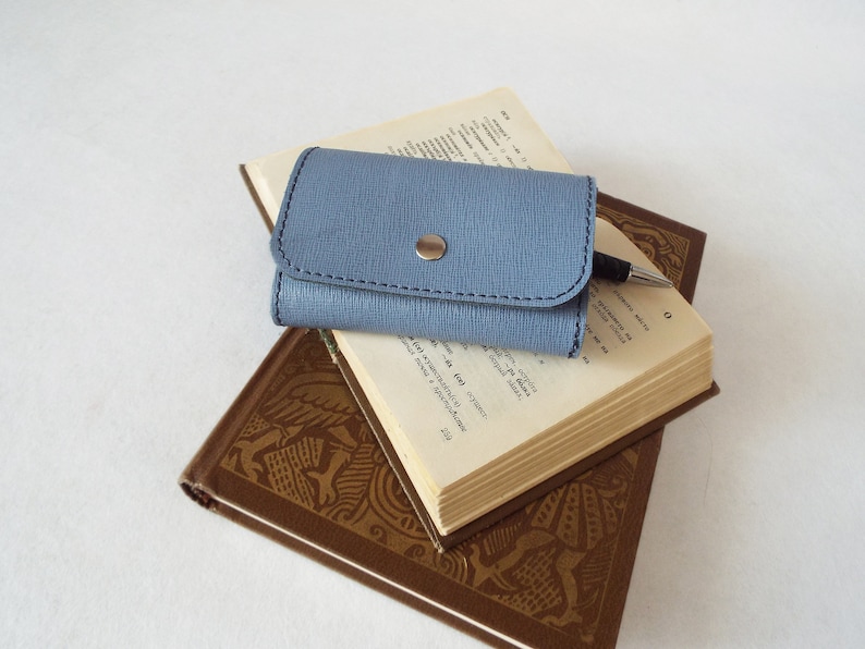 May include: A blue leather key holder with a silver button closure, resting on top of two books. The top book has a pen on it.