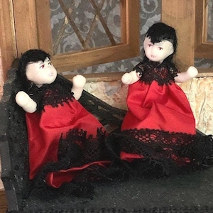May include: Two porcelain dolls with black hair and red dresses trimmed with black lace. The dolls are sitting on a black surface.