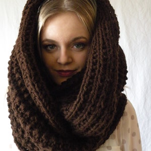 May include: A brown knitted infinity scarf made of thick yarn. The scarf is worn by a person with blonde hair and is folded over twice.