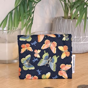 May include: A small, navy blue pouch with a zipper closure. The pouch is decorated with a repeating pattern of colorful butterflies and dragonflies. The pouch is sitting on a wooden surface.