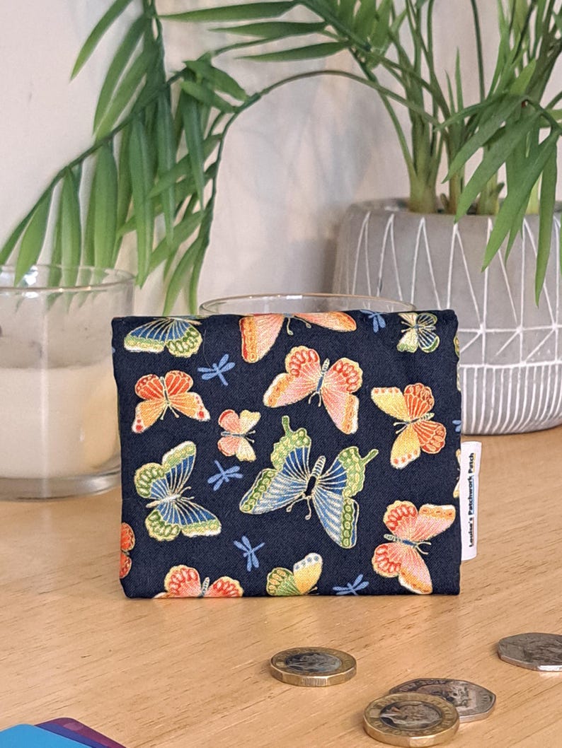 May include: A small, navy blue pouch with a zipper closure. The pouch is decorated with a repeating pattern of colorful butterflies and dragonflies. The pouch is sitting on a wooden surface.