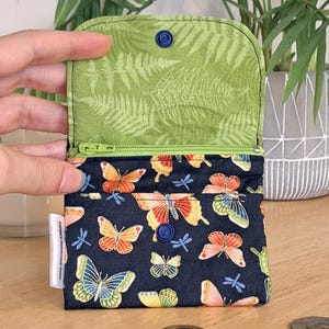 May include: A small zippered pouch with a dark blue background and a colorful butterfly print. The pouch is open and the inside lining is a light green with a fern pattern. The pouch has a blue button closure.