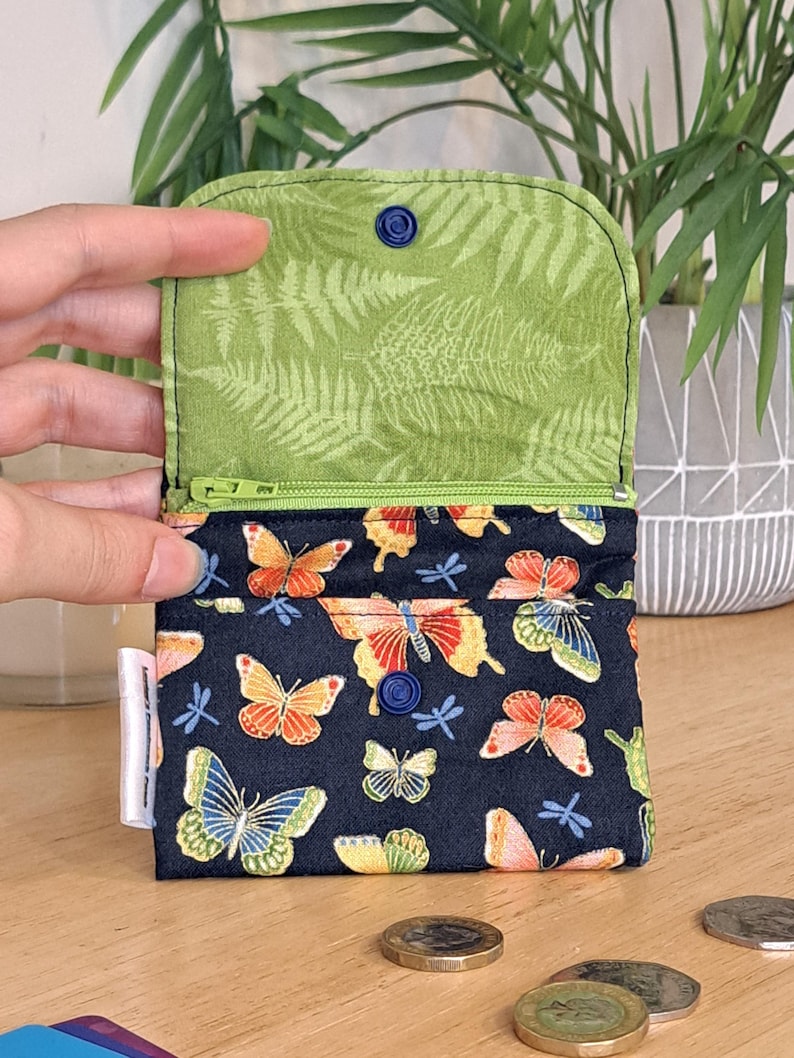 May include: A small zippered pouch with a dark blue background and a colorful butterfly print. The pouch is open and the inside lining is a light green with a fern pattern. The pouch has a blue button closure.