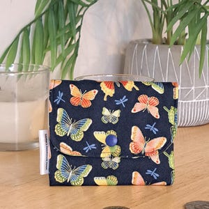 May include: A small black fabric wallet with a blue button closure. The wallet is decorated with a repeating pattern of colorful butterflies and dragonflies.