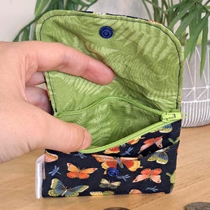 May include: A small zippered pouch with a green floral lining and a black exterior with a butterfly and dragonfly print. The pouch has a blue button closure.