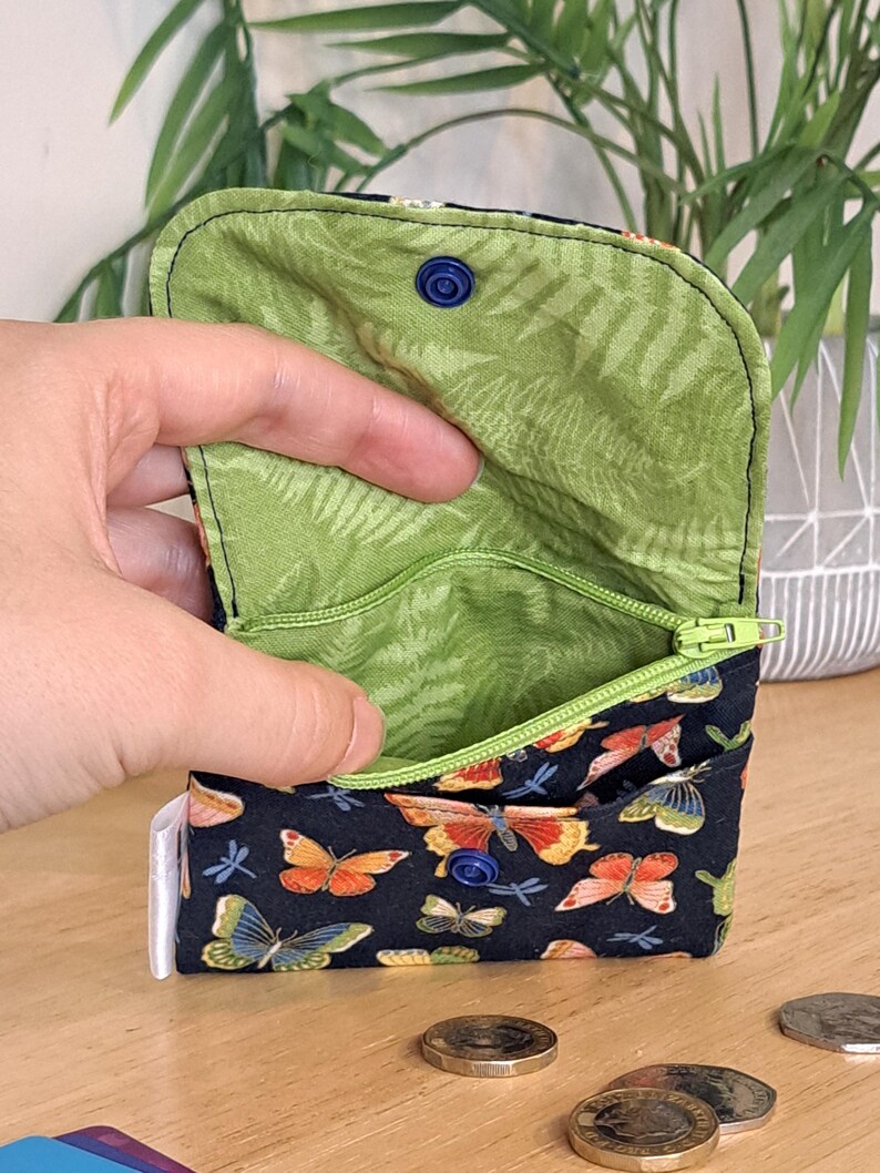 May include: A small zippered pouch with a green floral lining and a black exterior with a butterfly and dragonfly print. The pouch has a blue button closure.
