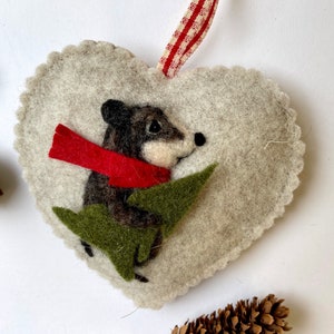 May include: A felt heart ornament with a brown mouse holding a green sprig of holly. The mouse is wearing a red scarf.