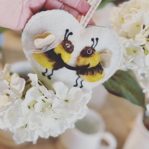May include: A white felt heart-shaped ornament with two embroidered bees, one yellow and one brown, with a yellow bow on top. The ornament is hanging from a white string.