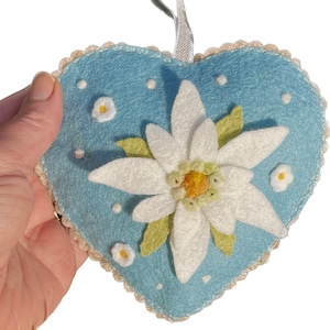 May include: A blue felt heart-shaped ornament featuring a white edelweiss flower and white dots. The ornament has a white ribbon loop for hanging.