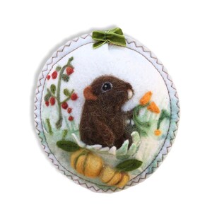 May include: A felt ornament featuring a brown mouse with a green bow, surrounded by green leaves, red berries, and yellow flowers.