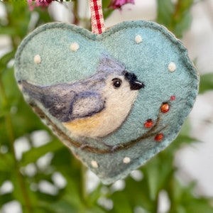 May include: A felt heart ornament with a bluebird perched on a branch with red berries. The background is a light blue with white dots.