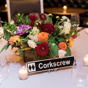 May include: A vibrant floral arrangement in a wooden box, featuring dahlias in shades of red, orange, and white, with greenery. A black and white sign that reads "Corkscrew" is in front of the flowers. A lit candle in a glass votive is in the foreground.