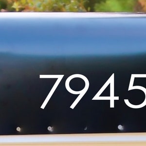 May include: A black mailbox with the white numbers 79452. The mailbox has a red flag and is mounted on a white base. The background is blurred with green and brown foliage.
