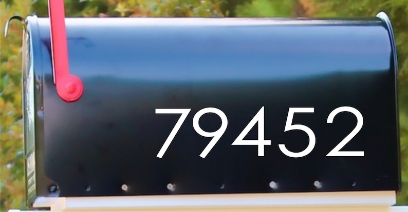 May include: A black mailbox with the white numbers 79452. The mailbox has a red flag and is mounted on a white base. The background is blurred with green and brown foliage.
