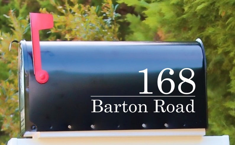 May include: A black mailbox with the address "168 Barton Road" in white lettering. The mailbox has a red flag on the side, and is set against a backdrop of green foliage.