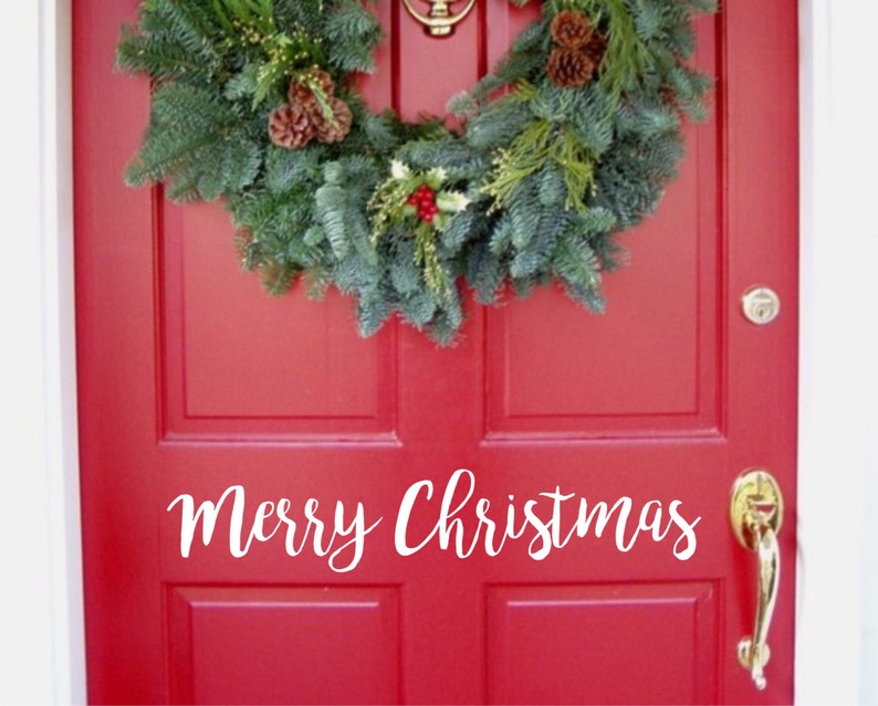 以下が含まれることがあります： 赤いドアにクリスマスのリースと白い筆記体で「Merry Christmas」の文字。リースは常緑樹の枝、松ぼっくり、赤い実でできています。金色のドアノブとノッカーが見えます。
