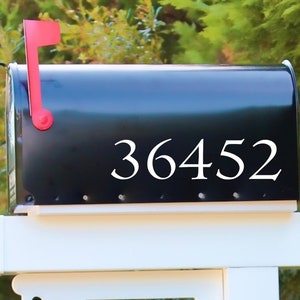May include: A black mailbox with the number 36452 in white lettering. The mailbox has a red flag and is mounted on a white post. Green foliage is visible in the background.