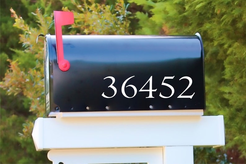 May include: A black mailbox with the number 36452 in white lettering. The mailbox has a red flag and is mounted on a white post. Green foliage is visible in the background.