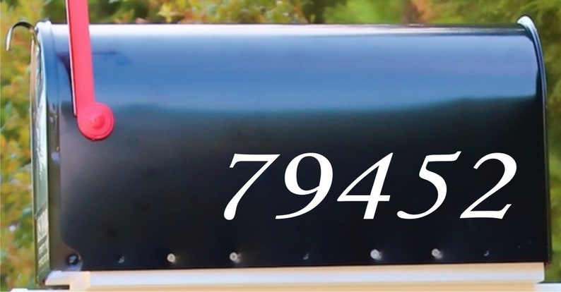 May include: A black mailbox with the white numbers 79452 displayed prominently on the side. The mailbox has a red flag and is set against a blurred green background.
