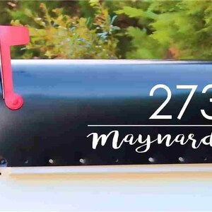 May include: A black mailbox with the address number 273 and the name "Maynard" in white lettering. The mailbox has a red flag and is set against a backdrop of green foliage.