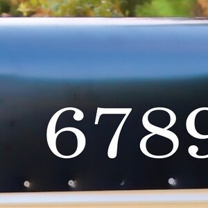 May include: A black mailbox with the white numbers 67890. The mailbox has a red flag and is mounted on a white base. The background is blurred green foliage.