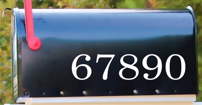 May include: A black mailbox with the white numbers 67890. The mailbox has a red flag and is mounted on a white base. The background is blurred green foliage.