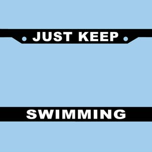 May include: Black license plate frame with the words "JUST KEEP SWIMMING" in white, set against a light blue background. The frame has a rectangular shape with rounded corners and is designed to fit a standard license plate.