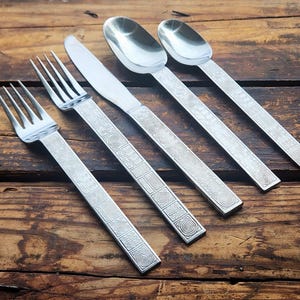 May include: Silverware set including two forks, a knife, and two spoons. The handles feature a repeating geometric pattern. The utensils are arranged diagonally on a rustic, wooden surface.