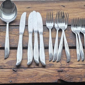 May include: A collection of vintage silverware, including spoons, forks, knives, and a ladle, arranged on a weathered wooden surface. The stainless steel utensils have a classic design, reflecting light. The arrangement suggests a formal dining setting.