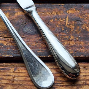 May include: Two silver utensils, a knife and a spoon, are lying on a wooden surface. The knife has a smooth, shiny handle and a sharp blade. The spoon has a rounded bowl and a long, slender handle.