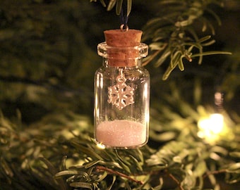 May include: A clear glass bottle with a cork stopper, filled with white glitter and a silver snowflake charm, hanging from a Christmas tree. The bottle is suspended by a blue string, creating a festive ornament.
