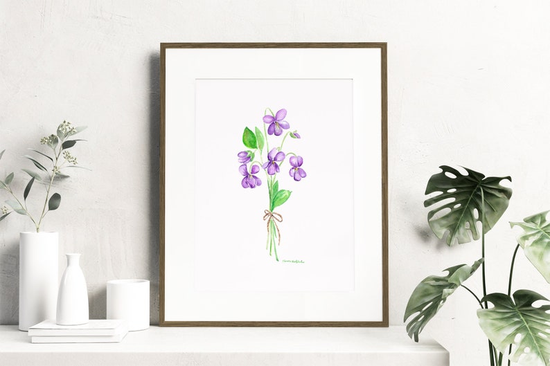 Puede incluir: Una pintura de acuarela de un ramo de violetas moradas con hojas verdes, atadas con un cordel. La pintura est&aacute; enmarcada en un marco de madera marr&oacute;n y se exhibe en un estante blanco.
