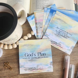 Puede incluir: Una composición de naturaleza muerta con una taza de café, tarjetas de acuarela con citas inspiradoras y bolígrafos. Las tarjetas tienen un diseño de acuarela pastel con el texto "God's Plan" y "The counsel of the Lord stands forever."