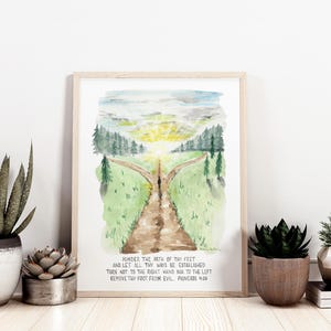 May include: Watercolor print in a light wood frame featuring a path splitting into two, leading to a bright sky. The artwork includes text from Proverbs 4:26. The print is displayed with potted succulents and a stack of books.