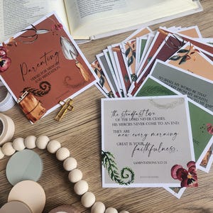 Pode incluir: Um conjunto de cartões com desenhos florais e texto, incluindo as palavras "Parenting" e "the steadfast love". Os cartões estão dispostos sobre uma superfície de madeira com uma cruz dourada, contas de madeira e um rolo de fita adesiva.