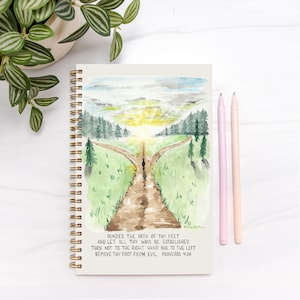 May include: Spiral-bound notebook with a watercolor painting of a path splitting in two, leading to a bright sunset. The notebook has gold spiral binding and the text "PONDER THE PATH OF THY FEET...PROVERBS 4:26". Two pens, pink and peach, are next to the notebook.