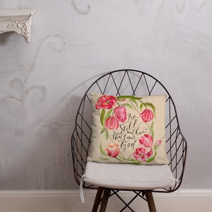 May include: A beige throw pillow with a floral design featuring pink tulips and a gold geometric frame. The pillow has the text "Be Still and know that I am God" in black calligraphy.