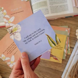 May include: A hand holding a light blue card with text from Psalm 91:2, surrounded by other cards and a book. The cards feature floral illustrations and text. The background includes a Bible and decorative elements.